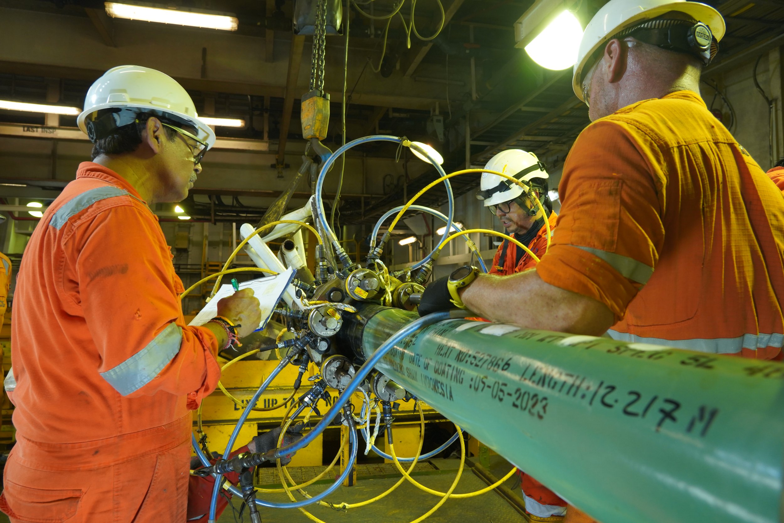 Workers in orange uniforms and white helmets working on a large mechanical component with numerous tubes and tools in an industrial setting.