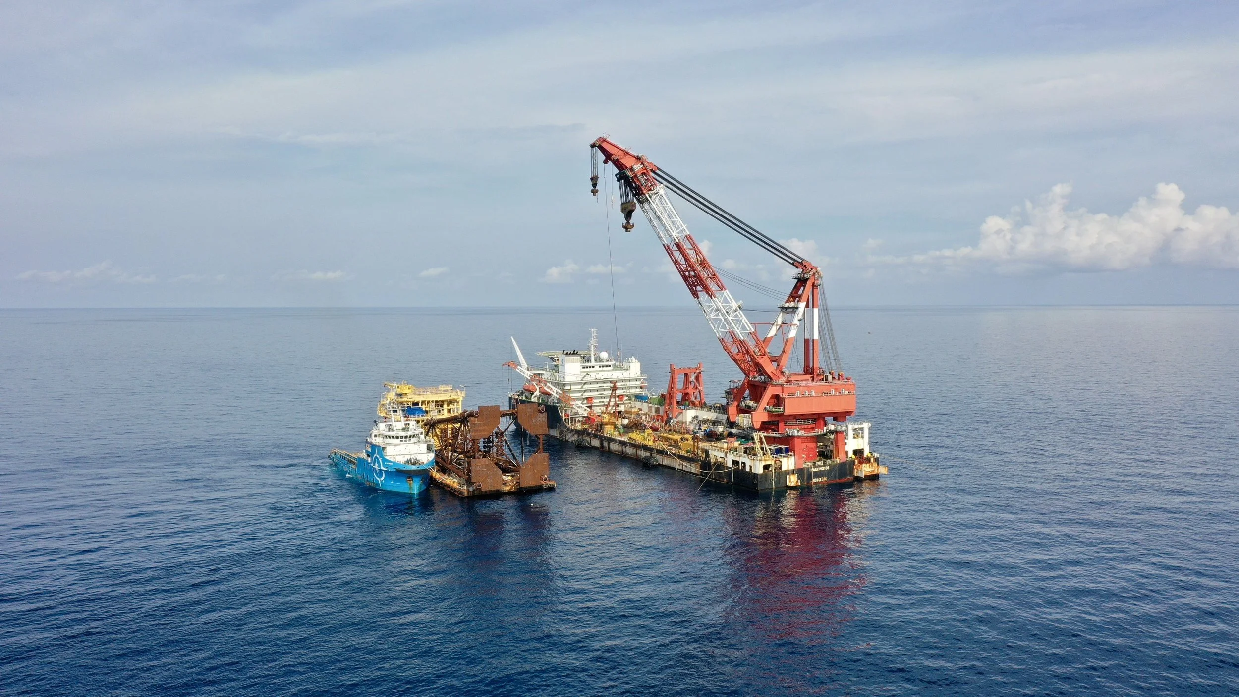 Offshore oil drilling rig with cranes and support vessels on calm ocean water under a partly cloudy sky.