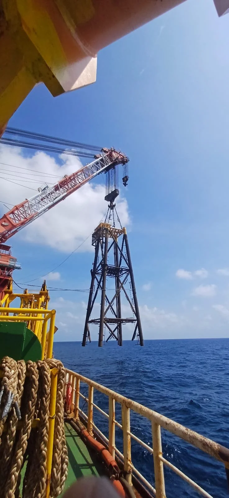 A crane lifting equipment on an offshore oil rig platform extended over the water, with a blue sky and some clouds in the background.