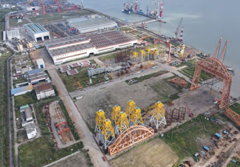 Aerial view of a construction site with cranes, industrial buildings, and a nearby body of water.