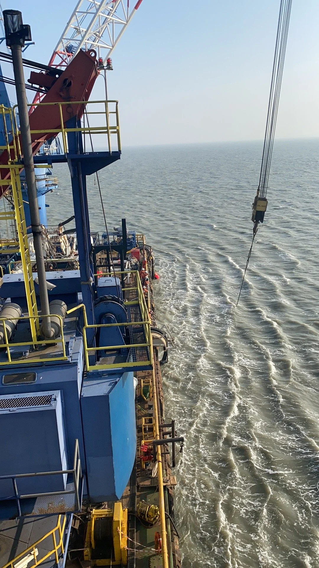 View of a large offshore oil platform with equipment, railings, and a crane, floating in the ocean with visible waves and a clear sky.