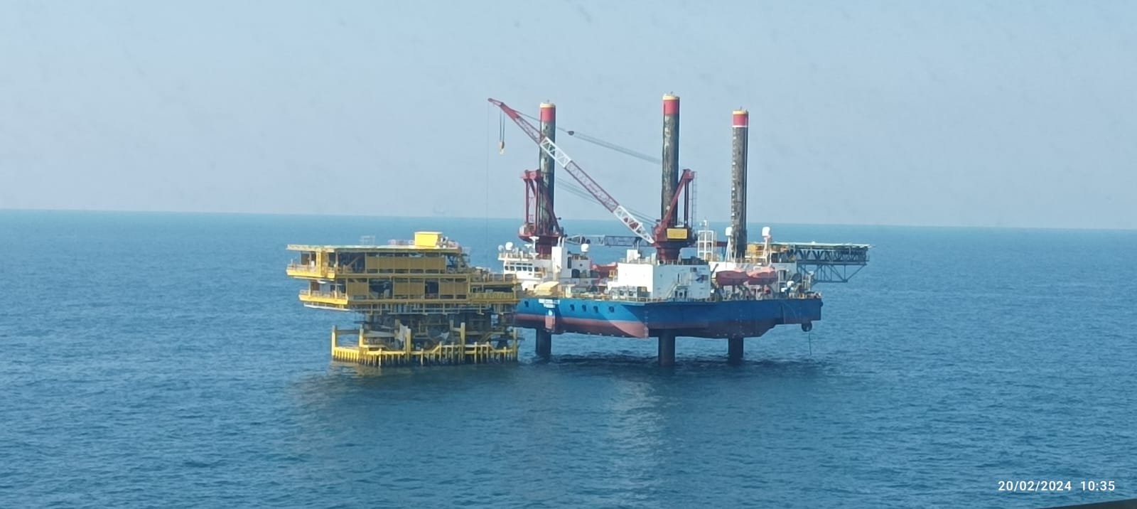 An offshore oil rig floating in the ocean with a clear sky in the background. The rig has a yellow platform and three tall derrick structures. The date and time are displayed in the bottom right corner as 20/02/2024 10:35.
