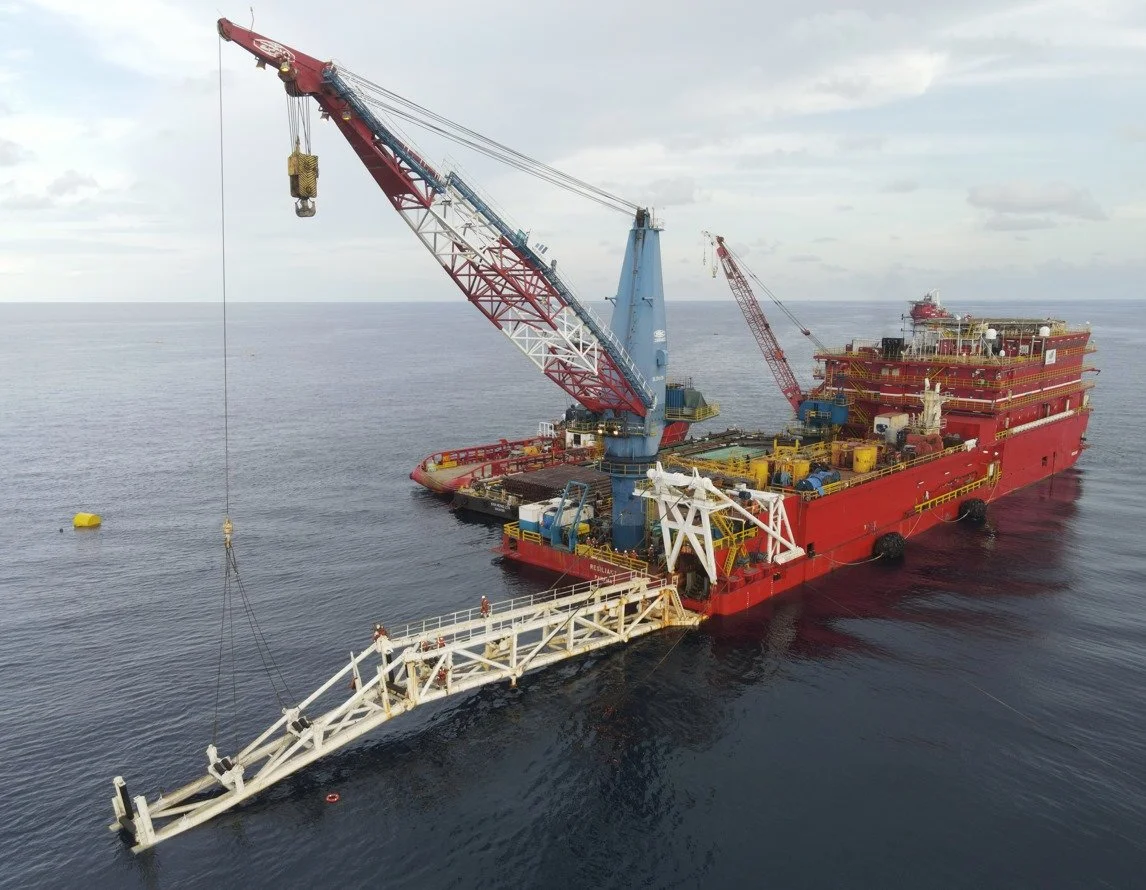 A large offshore drilling platform and crane vessel floating on the ocean, equipped with cranes, pipelines, and various industrial equipment for oil or gas extraction.