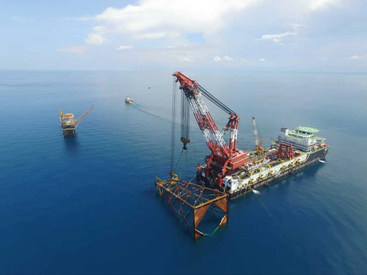 A large offshore construction vessel with a crane installing a bridge or platform structure in the ocean, with a smaller boat nearby and another platform in the background.