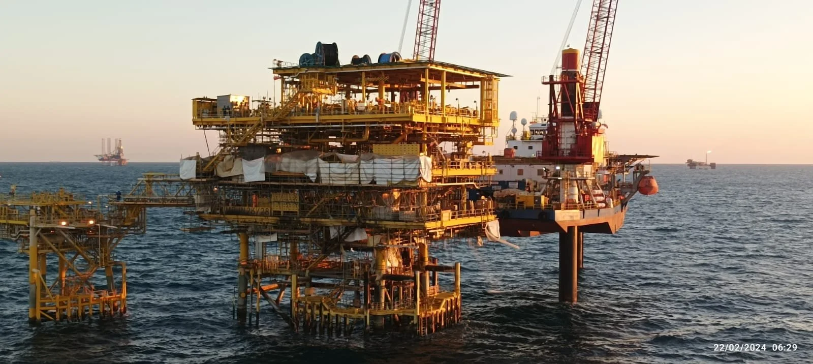 Offshore oil drilling rig at sea during sunset with two oil tankers in the background.