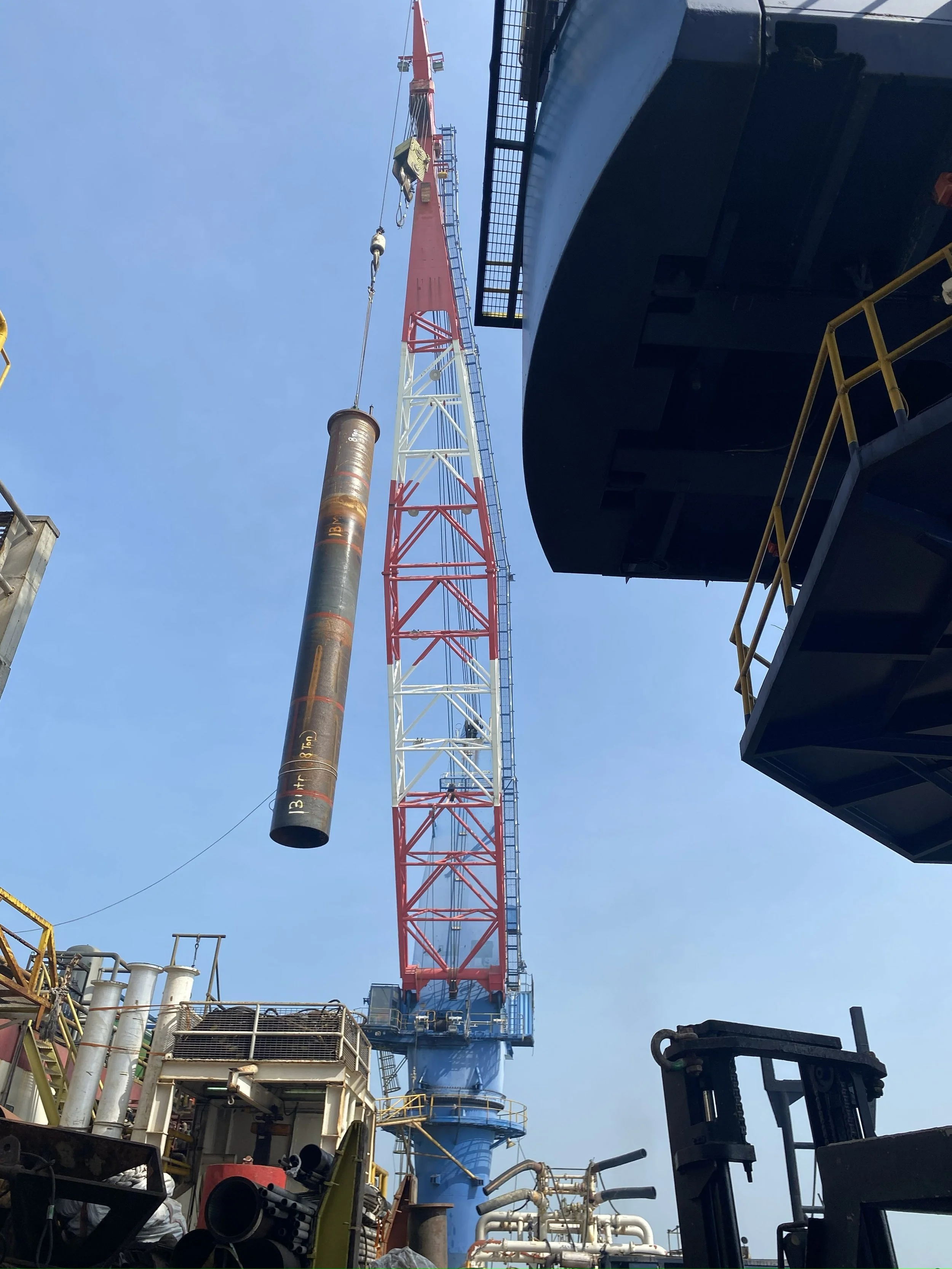 Construction site with a large crane lifting a heavy pipe into the air, surrounded by industrial equipment and structures.