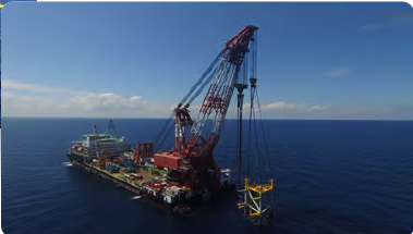 A large offshore oil drilling rig with a crane on a floating platform at sea, under a blue sky.