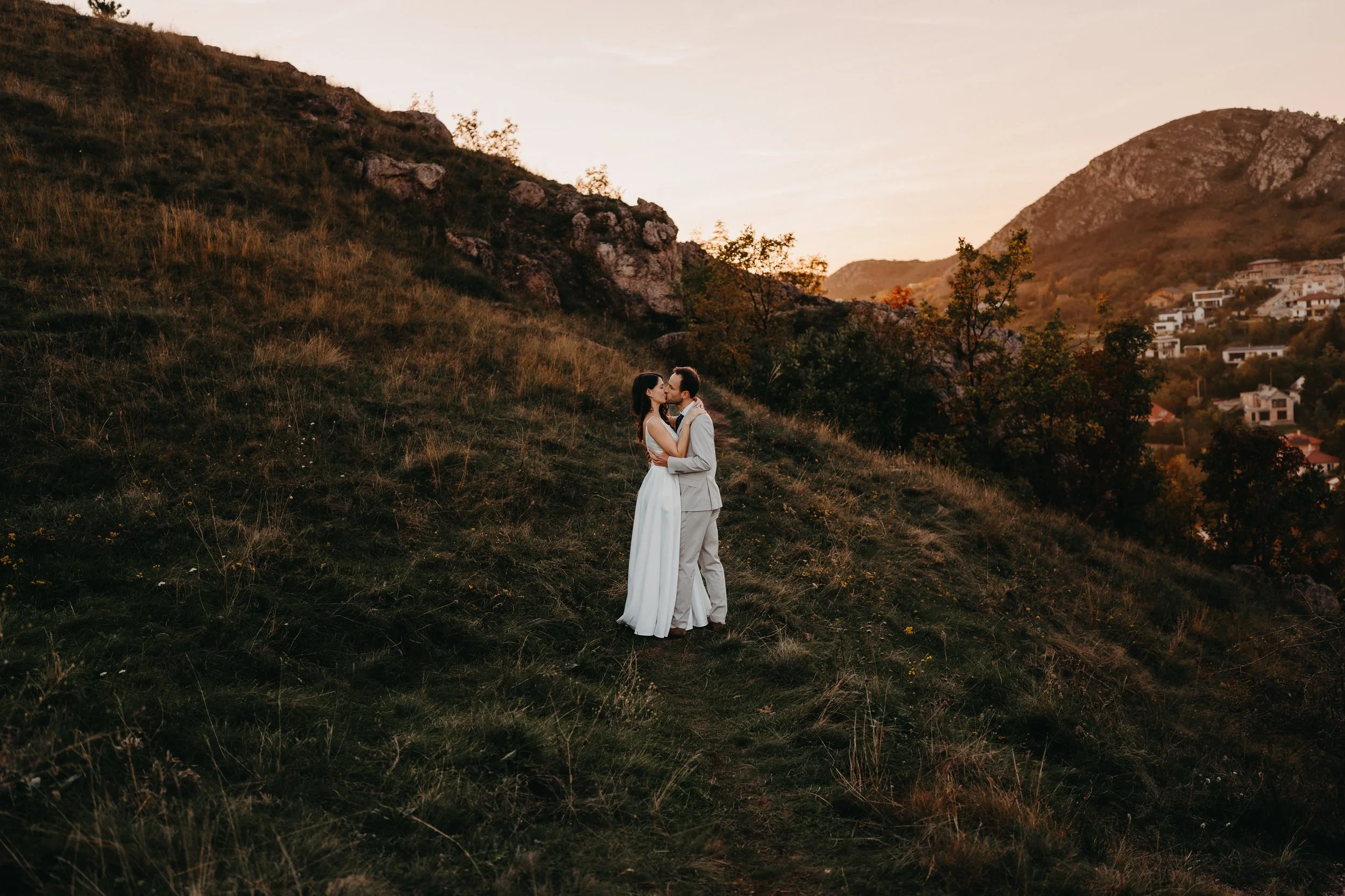 A couple in wedding attire sharing a kiss on a hillside at sunset with hills and houses in the background.