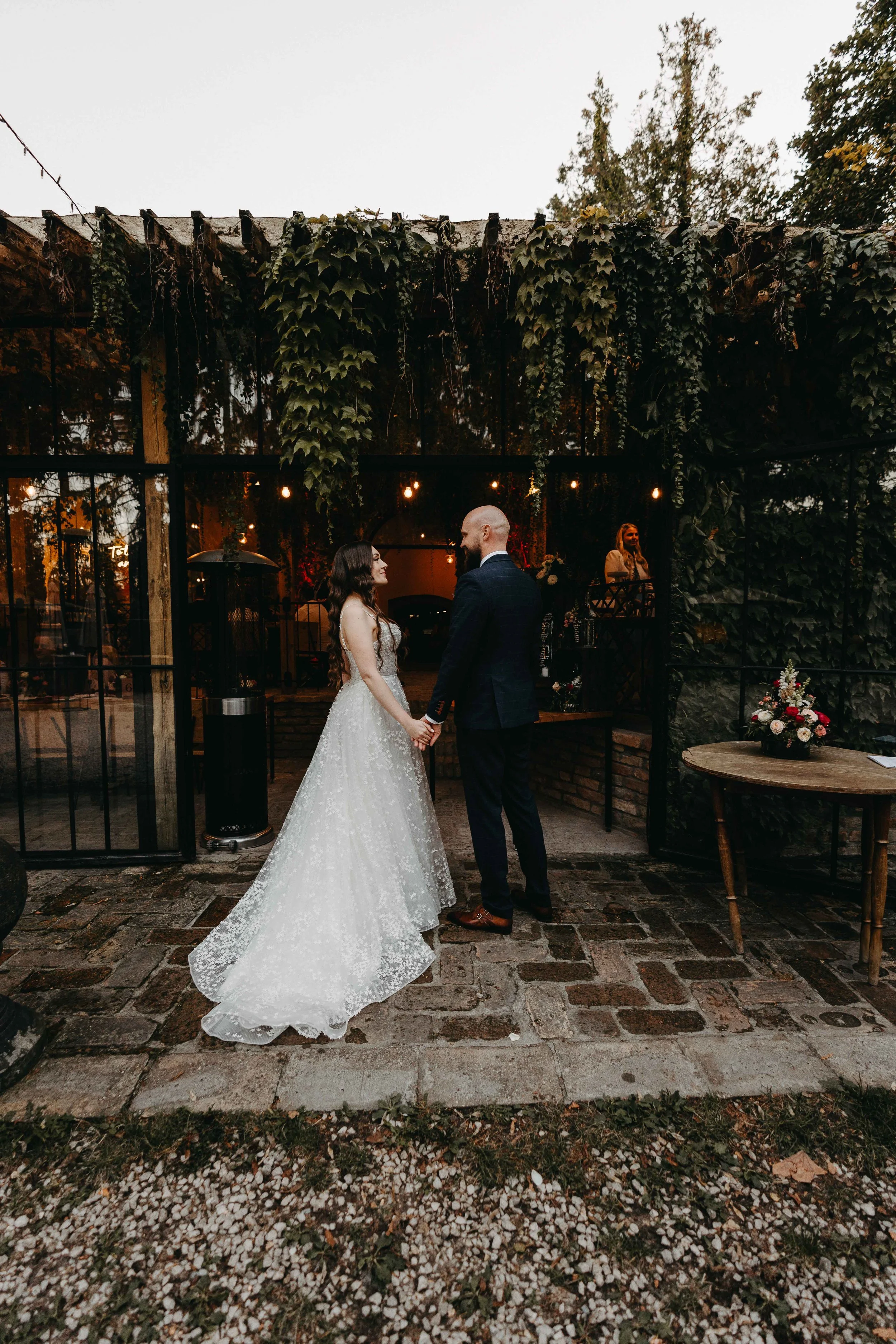 A bride and groom holding hands and facing each other during their wedding ceremony outside at dusk, with a glass wall and greenery in the background.