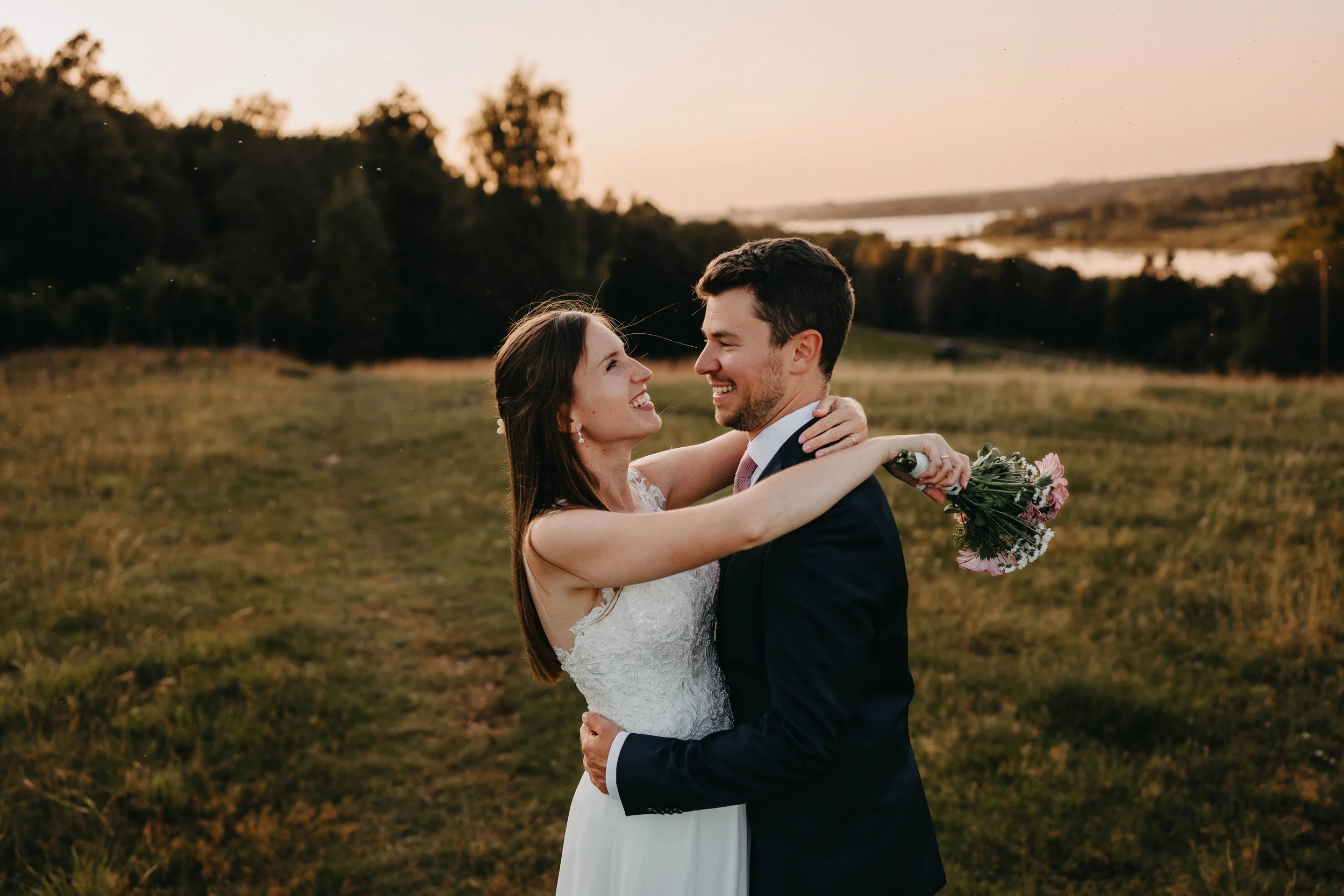 A bride and groom embrace outdoors at sunset, with the bride holding a bouquet of pink and white flowers, standing in a grassy field with trees and a river in the background.