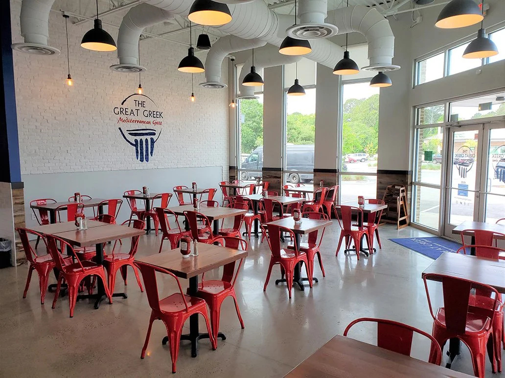 Interior of a Greek restaurant with white brick walls, black pendant lamps, red metal chairs, and light wood tables. Large windows allow natural light, and the logo of the restaurant, 'The Great Greek Mediterranean Grill,' is painted on the wall.