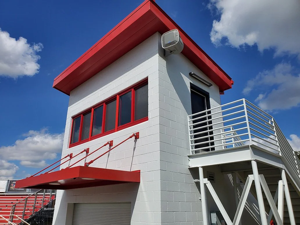 Exterior of a white building with red accents, large windows, metal stairs, and a rooftop area, under a partly cloudy sky.