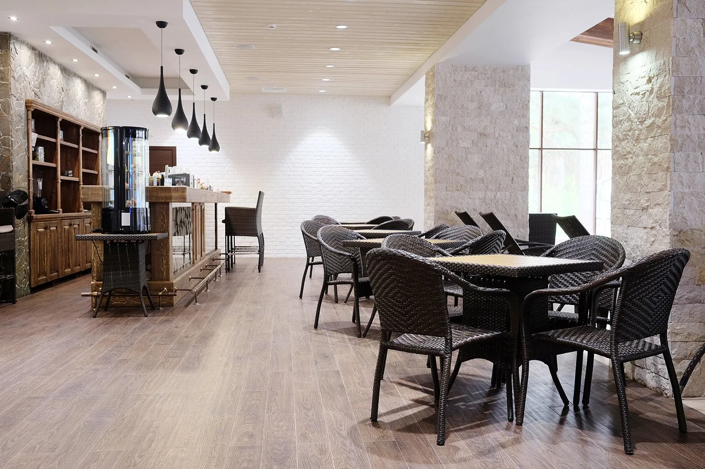 Interior of a modern cafe or restaurant with wicker chairs and tables on hardwood floors, a white brick wall, large window, and a wooden bar area with pendant lights.