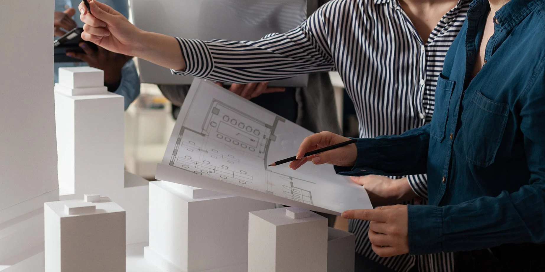 People working on an architectural model and blueprint. One person is holding and pointing at the blueprint with a pen, while others are observing. The scene appears to be in a design or planning studio.