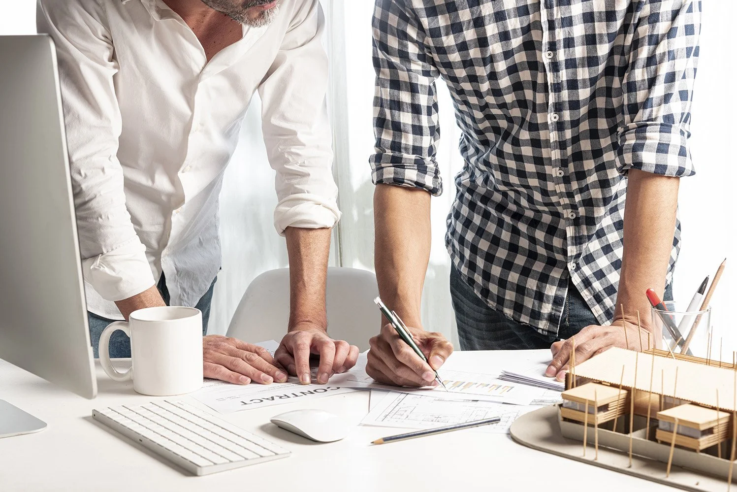 Two people are working on architectural plans at a desk, one is writing while the other is pointing at the plans, with a model of a building and a computer nearby.