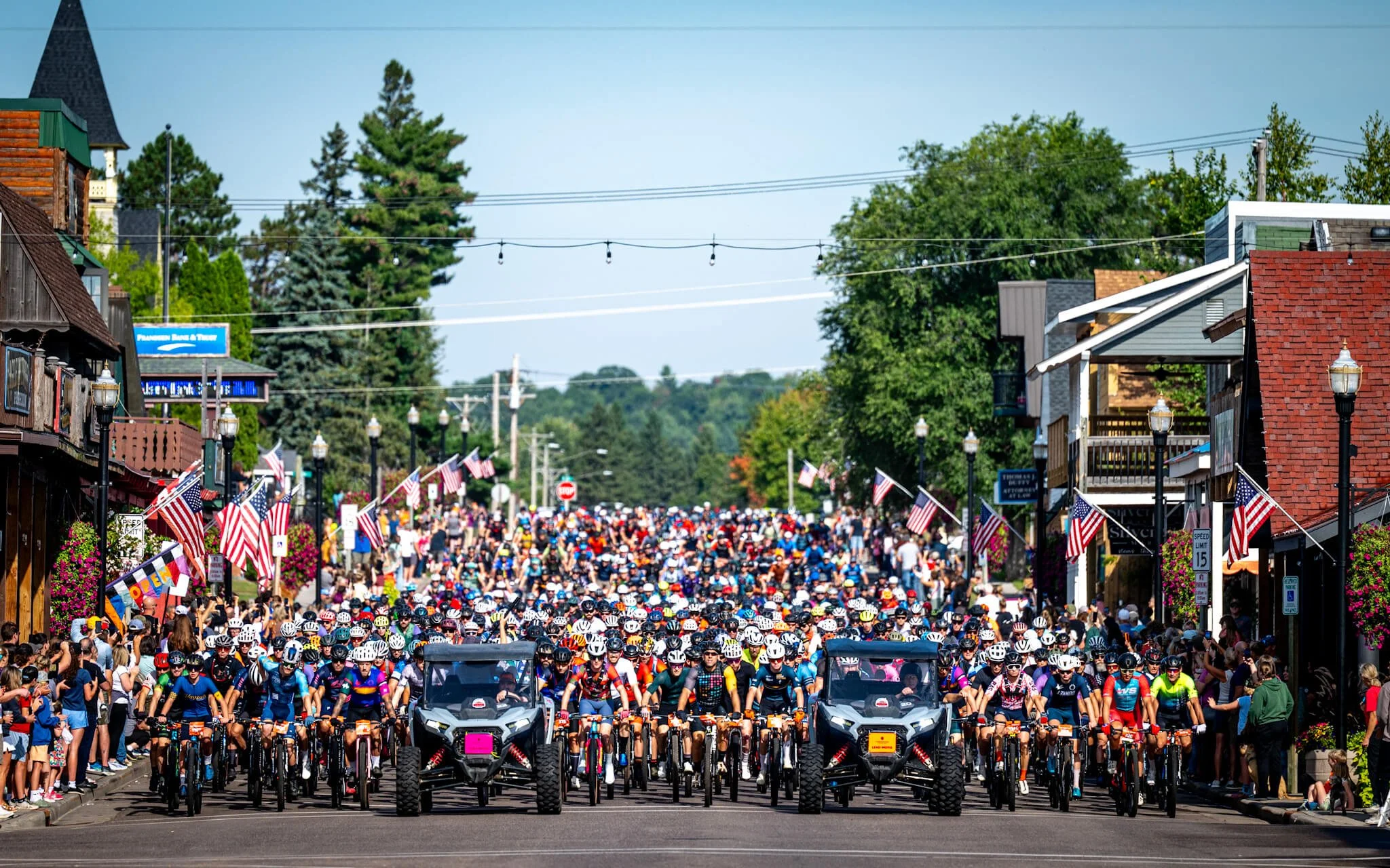A large group of cyclists at the start of a race on a patriotic-themed street, with flags and spectators on both sides.