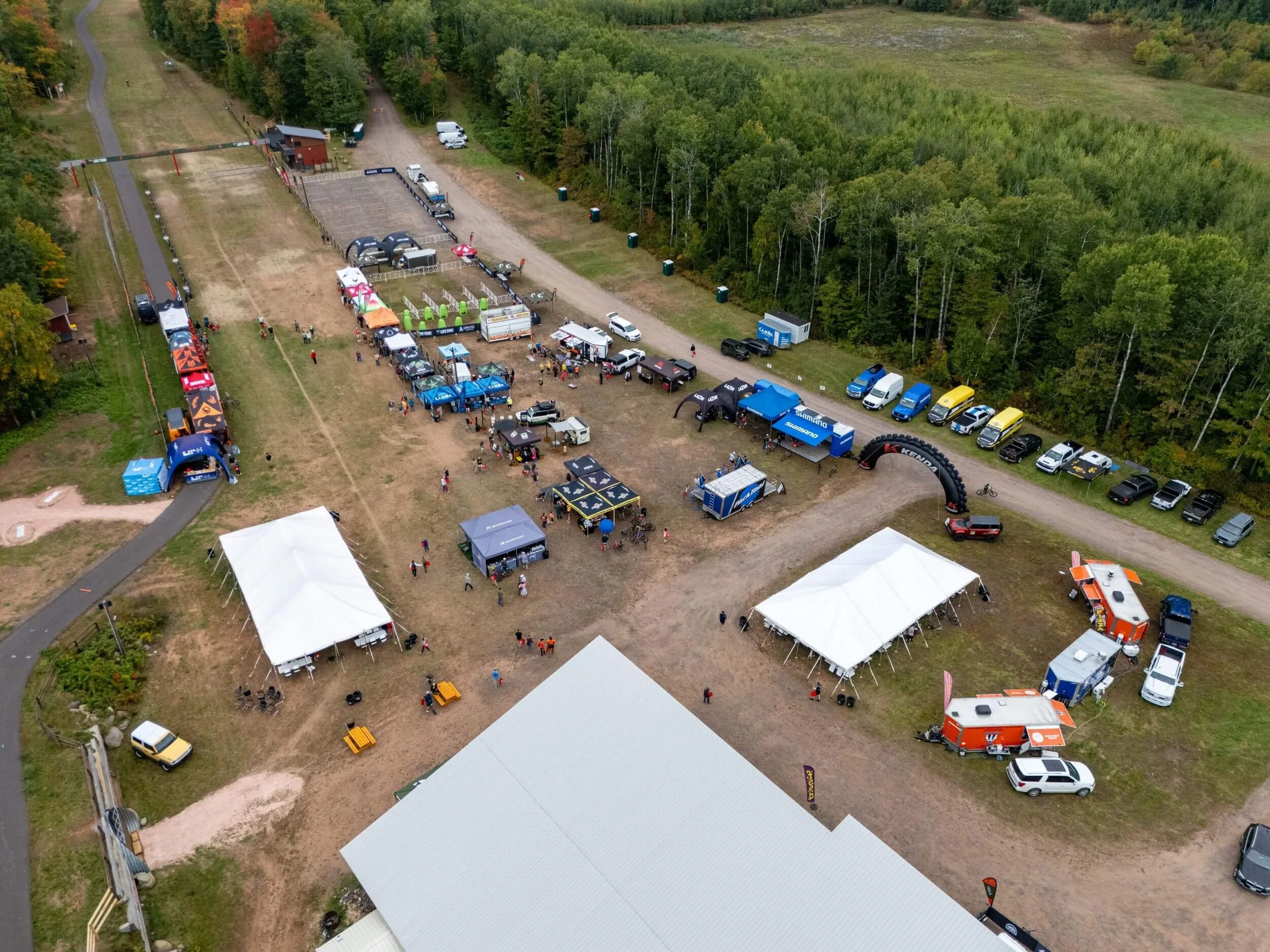 An aerial view of an outdoor event with tents, food trucks, and vehicles on a grassy field bordered by trees.