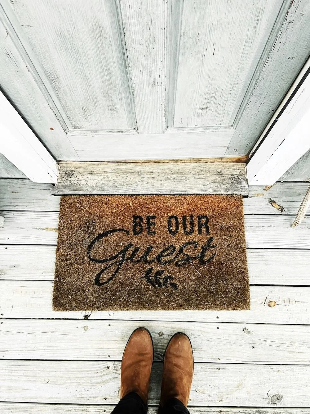 A welcome mat outside a wooden door with the words 'Be Our Guest' written on it, and a pair of brown boots visible at the bottom of the image.