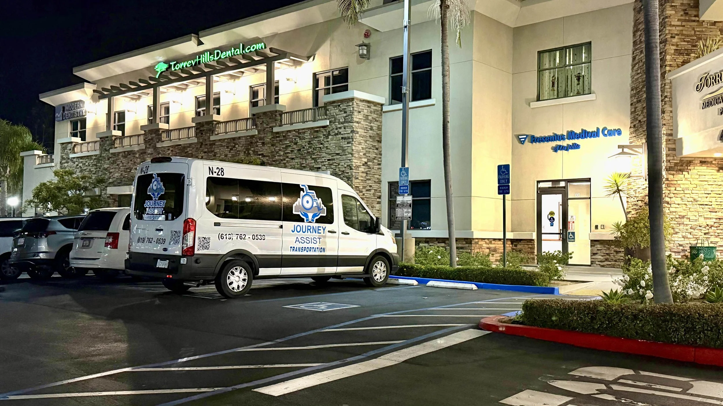 Nighttime view of a two-story medical building with signs reading "Torrey Hills Dental" and "Presentus Medical Care". In front of the building, there is a parking lot with several parked cars, including a white van labeled "Journey Assist Transportation". The building has exterior lighting, landscaping with bushes, trees, and white flowers, and designated handicapped parking spots.