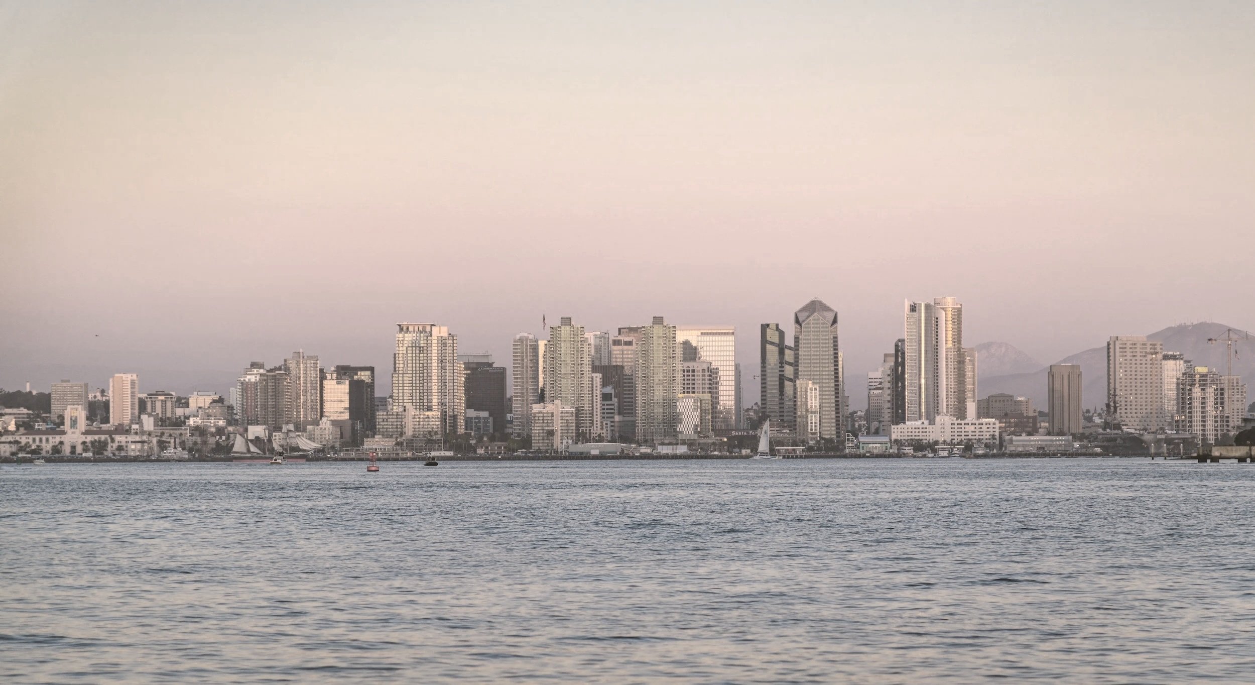 City skyline with tall buildings along the waterfront, sailboats on the water, and mountains in the background during dusk or early evening.