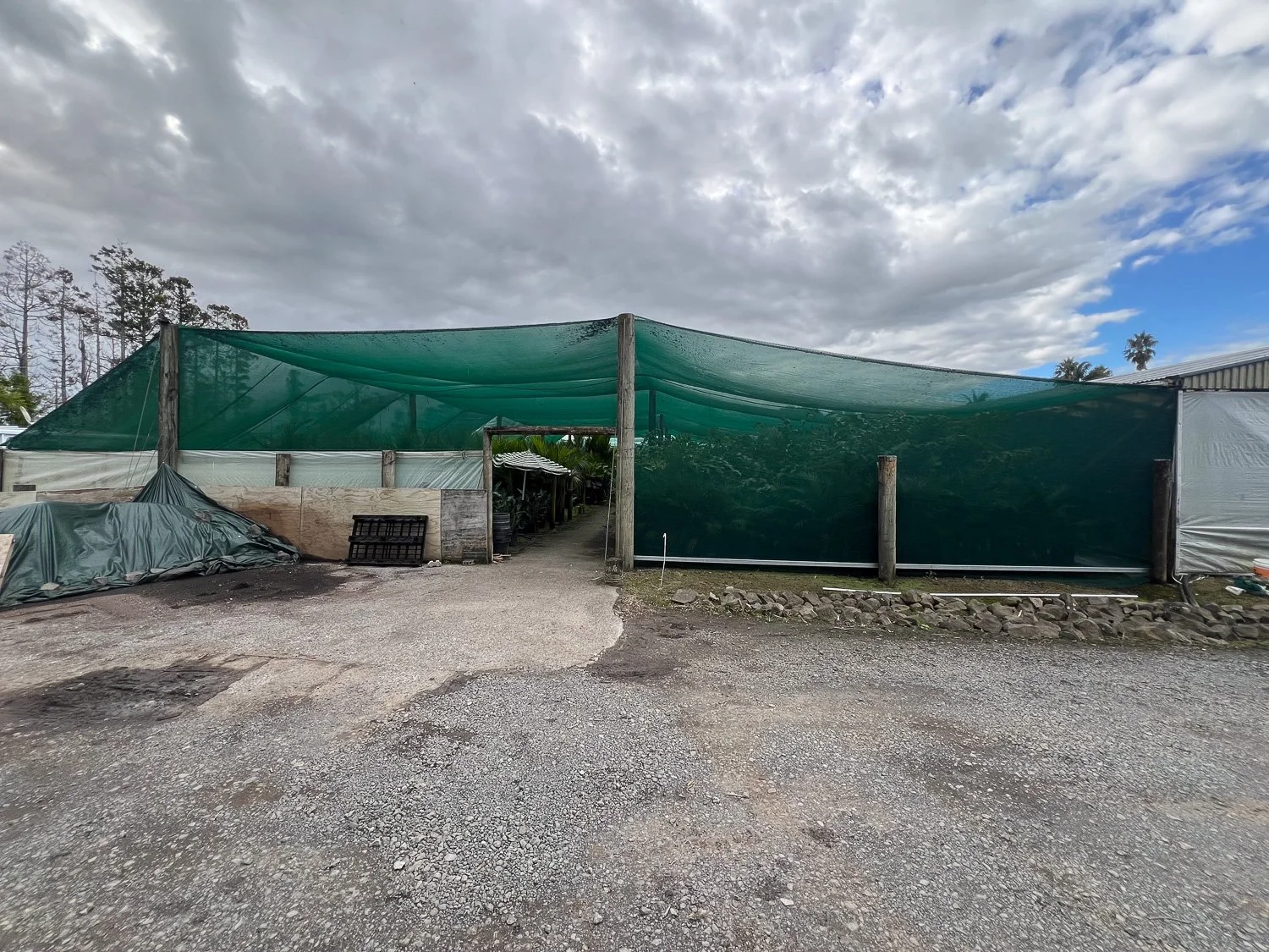 An outdoor area with a green shade cloth covering a garden or plant nursery, gravel ground, and cloudy sky.