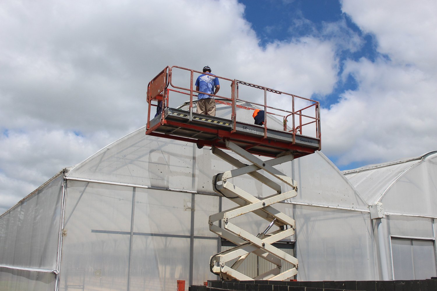 Workers on a red elevated lift working on a greenhouse with white plastic cover, set against partly cloudy sky.