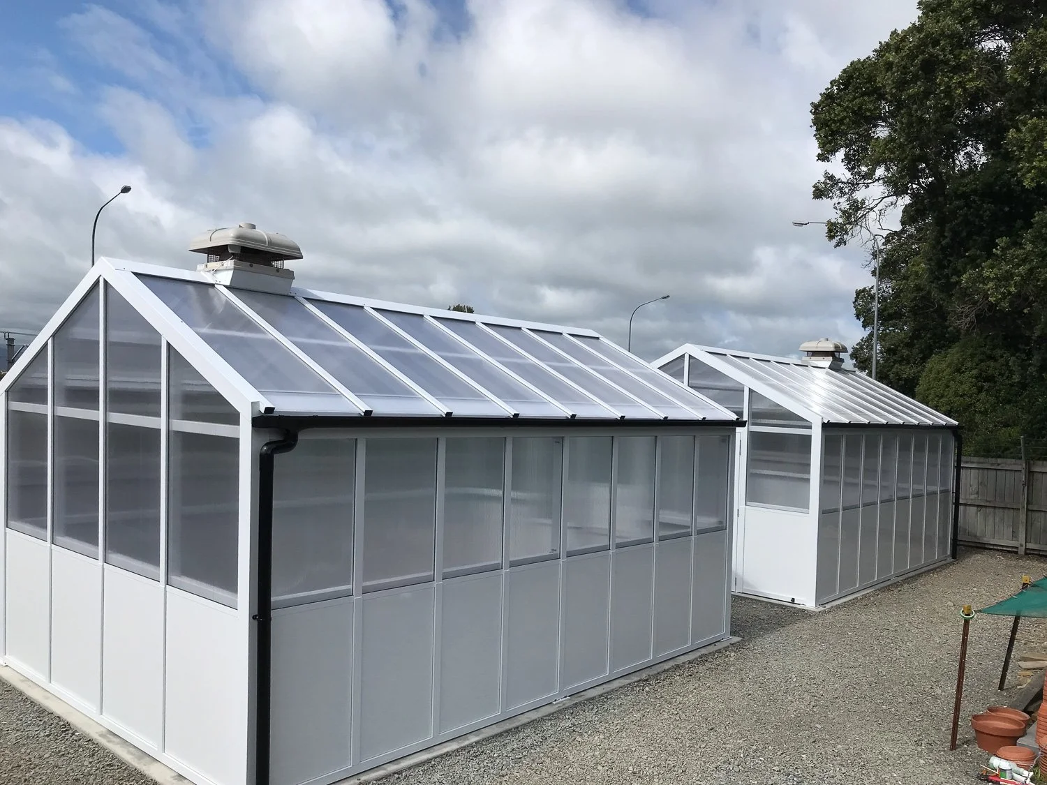 Two white metal-framed greenhouses with transparent panels, situated outdoors on a gravel surface, with one greenhouse in the foreground and the other slightly behind. There are trees and a cloudy sky in the background.