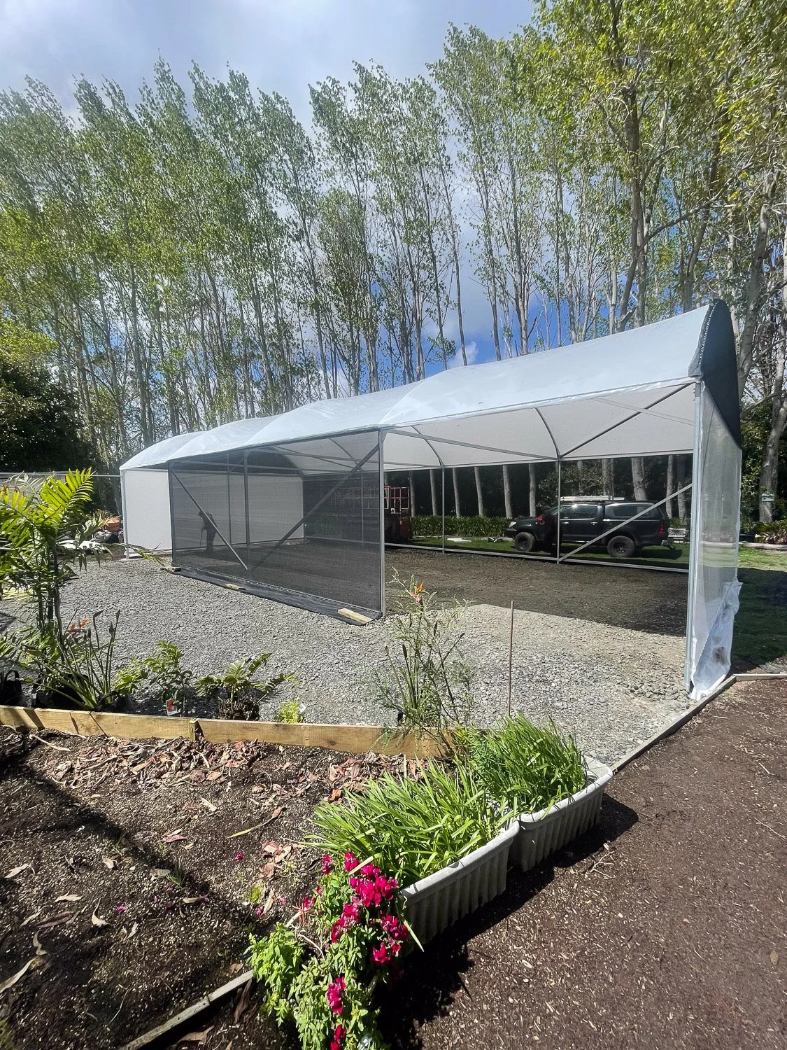 A white outdoor greenhouse with a black mesh window on one side, situated on a gravel surface next to a garden bed with flowering plants, with trees and parked cars in the background under a clear sky.