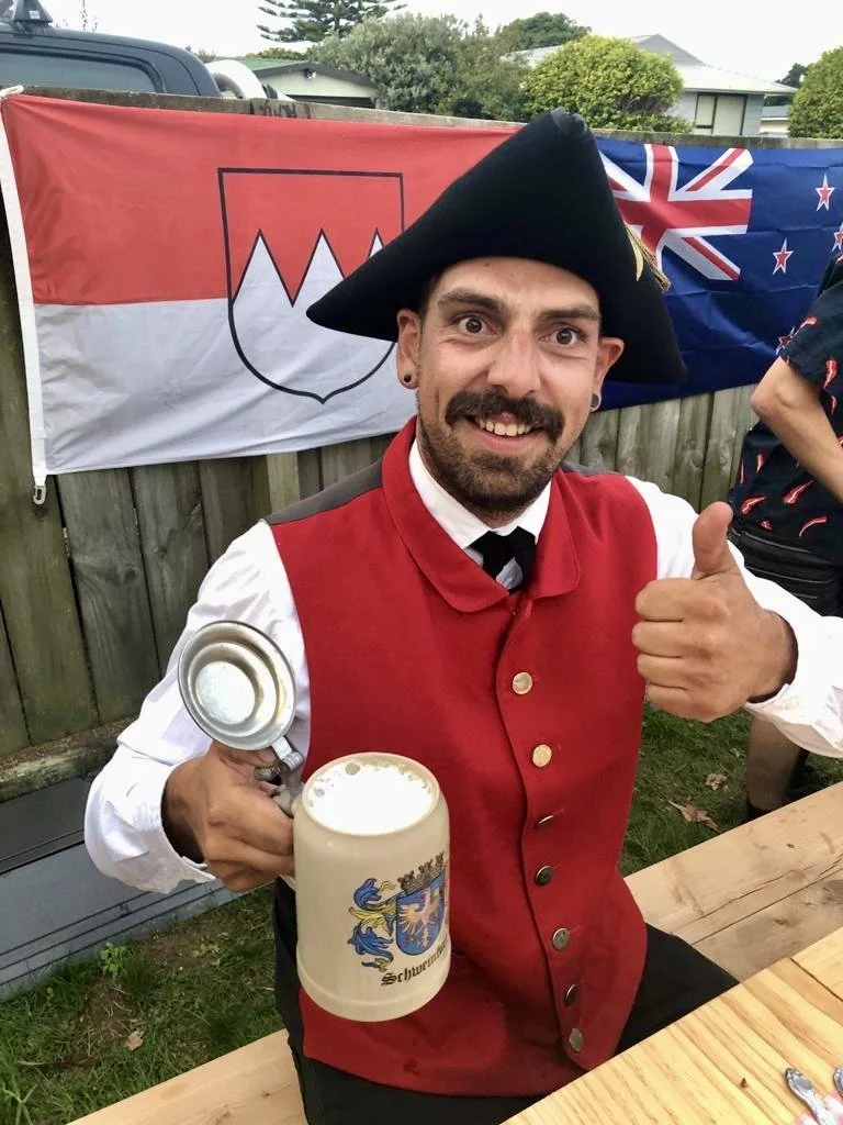 Man dressed in historical costume, wearing a tricorn hat, red vest, and white shirt, holding a beer stein with a crest, giving a thumbs up, with flags in the background.