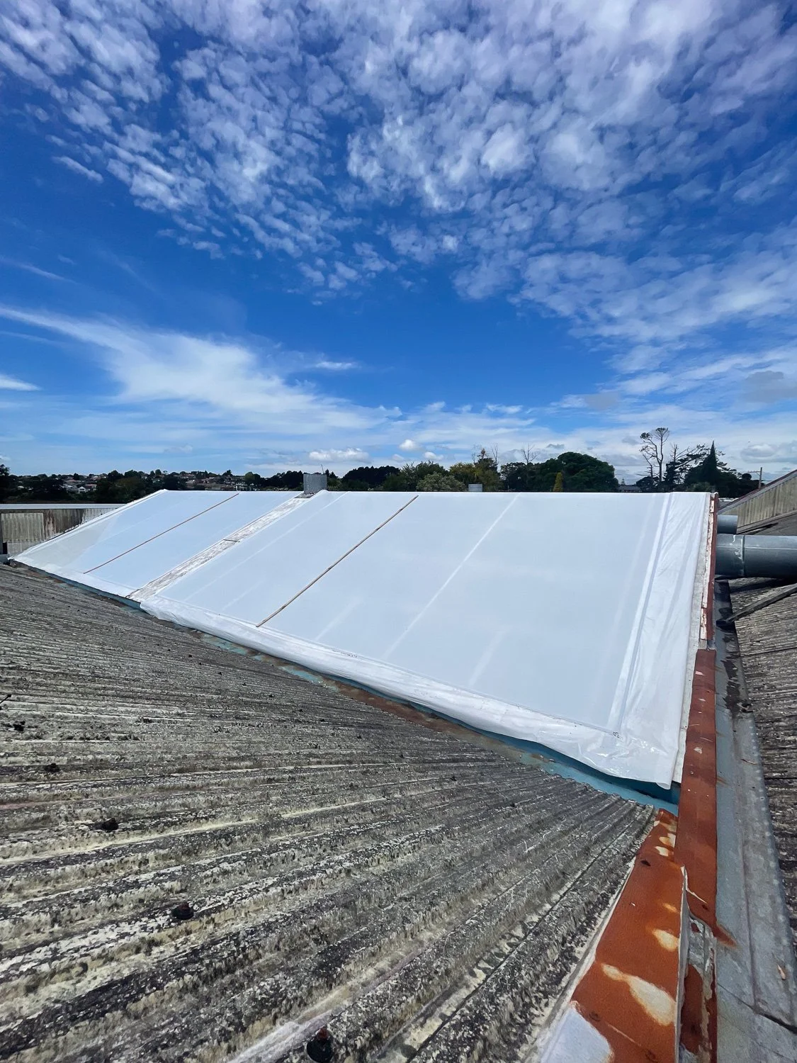 Roof with new white insulation or weatherproofing material installed on the upper section, contrasting with older, weathered gray roofing on the lower section. A cloudy blue sky is visible above.
