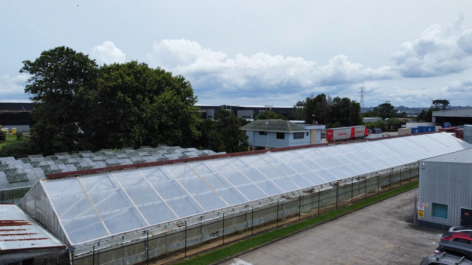 A large greenhouse with transparent panels situated on a fenced outdoor area in an industrial zone under a cloudy sky, with trees and industrial buildings in the background.