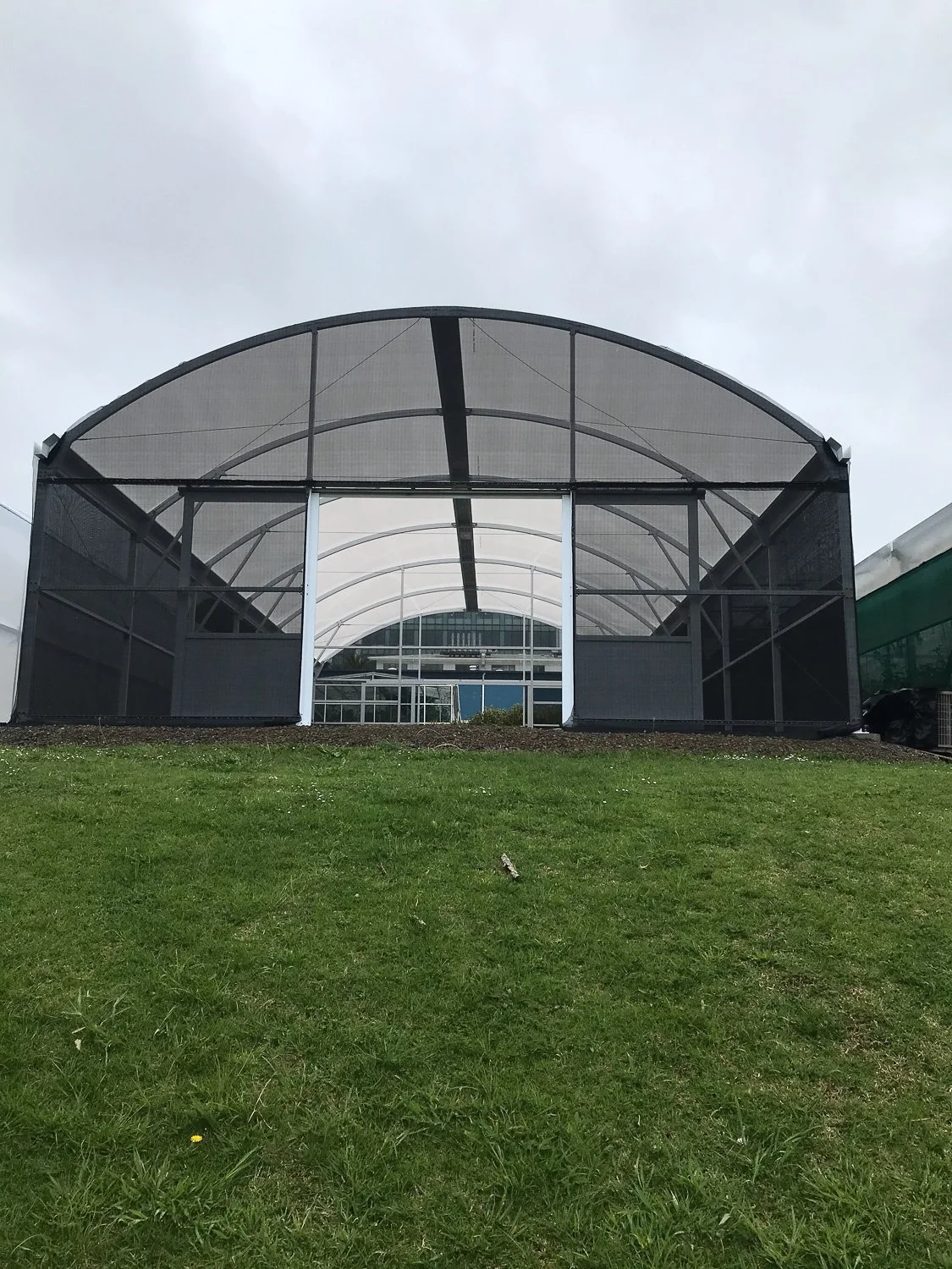 Green grassy lawn in front of a large black and white greenhouse structure with a curved roof and glass walls under a cloudy sky.
