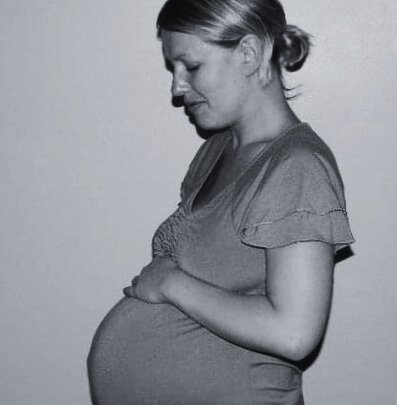 A pregnant woman with her eyes closed, gently holding her belly with both hands, standing sideways against a plain background.