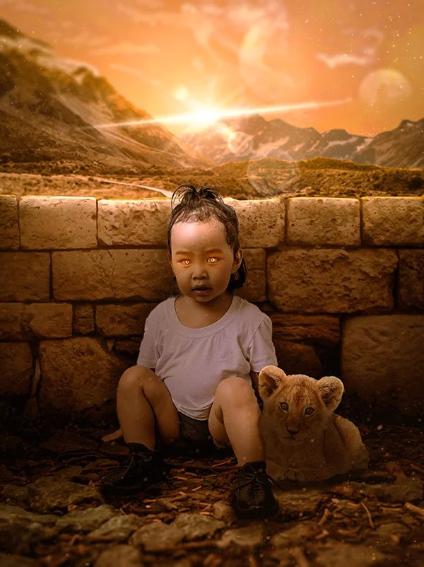 A young child with glowing eyes sits on rocky ground in front of a stone wall, holding a lion cub; a sunset and mountainous landscape with a bright sun and lens flares is visible in the background.