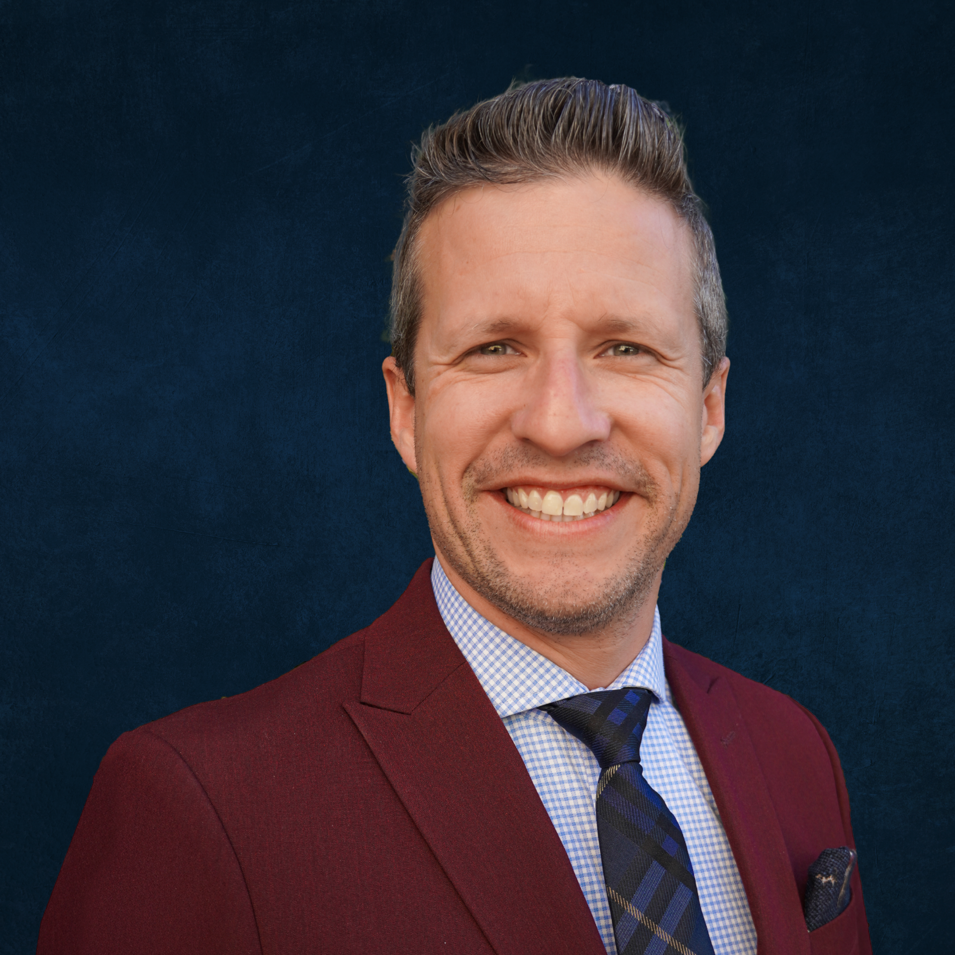 A smiling man with short, styled hair wearing a burgundy suit, a checkered shirt, a patterned tie, and a pocket square, posed against a blue background.