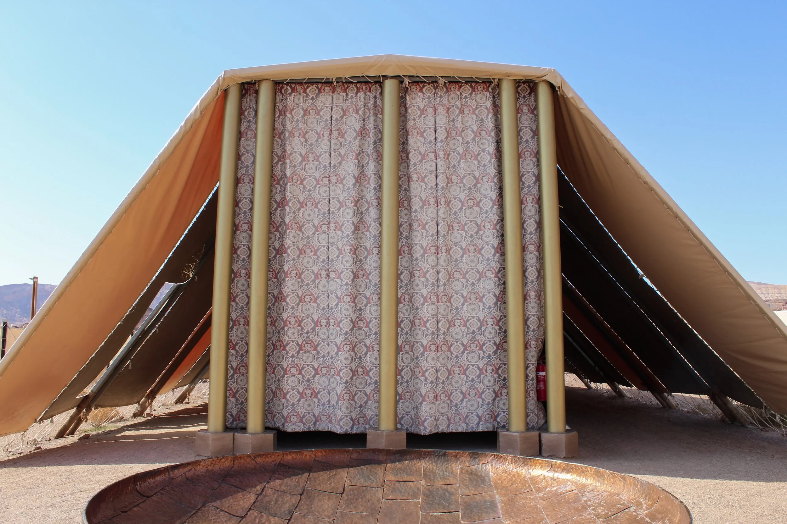 A large triangular tent with vertical wooden panels and beige fabric sides, set in a desert landscape with mountains in the background, under a partly cloudy sky, and a makeshift barrier surrounding the area.