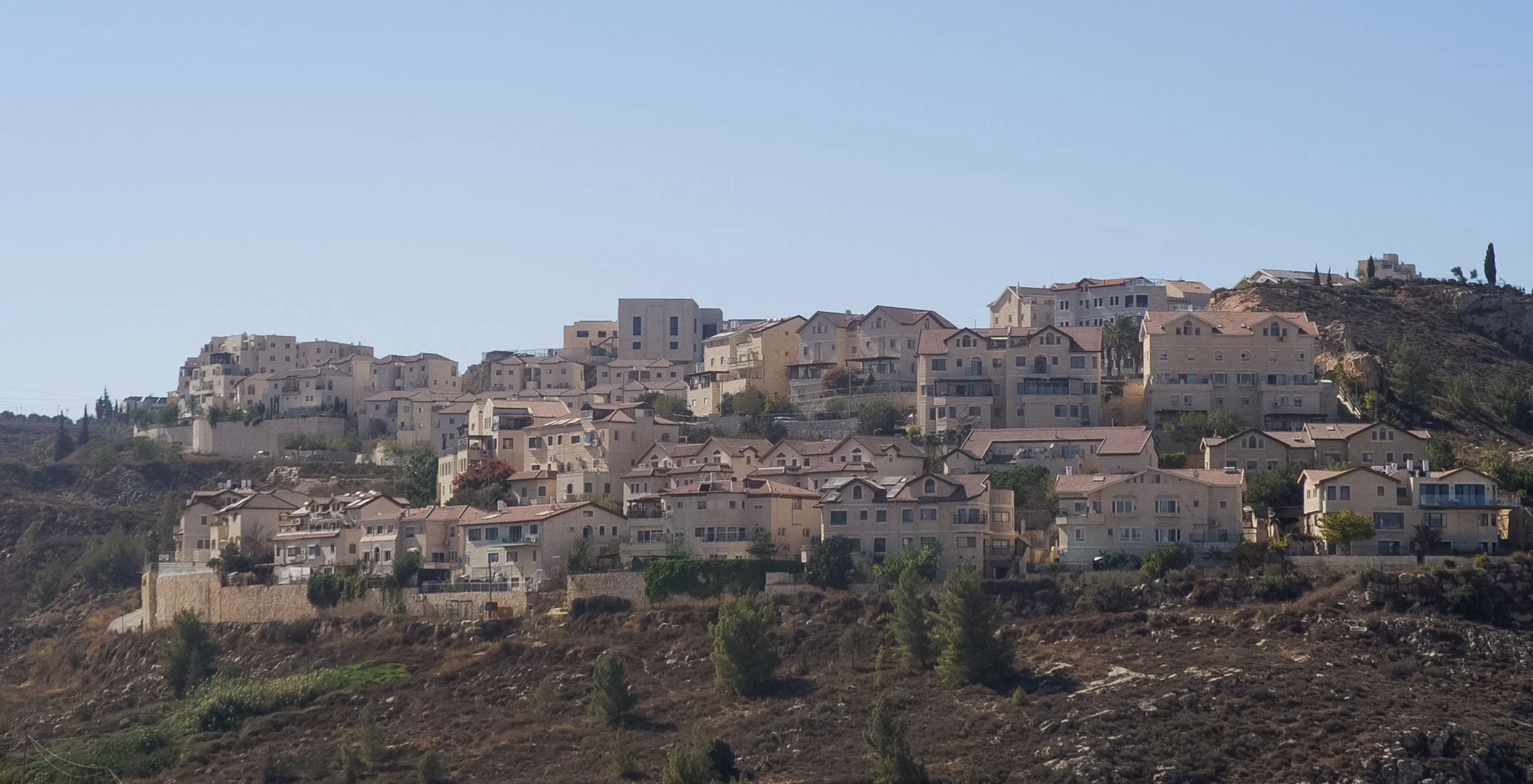 Hilly residential neighborhood with numerous multi-story houses with light-colored exteriors and red roofs, situated on a hillside under a clear blue sky.