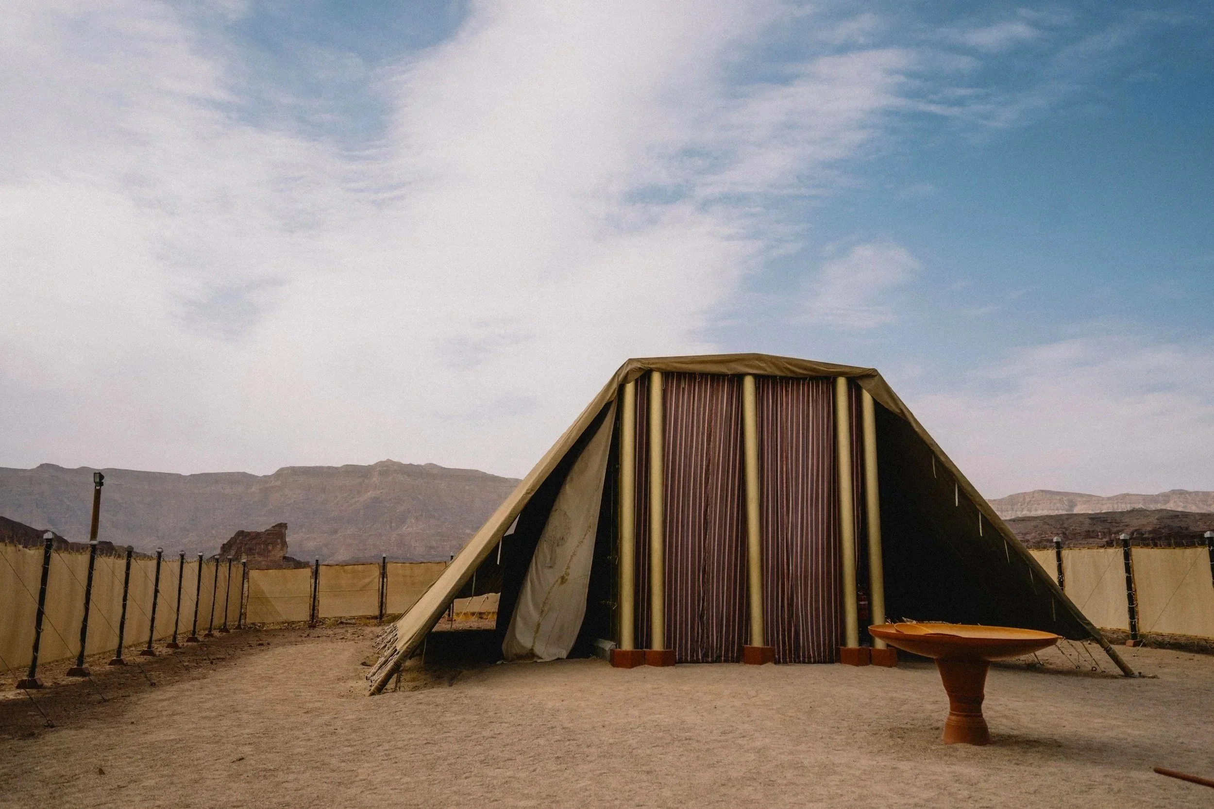 A large triangular tent with vertical wooden panels and beige fabric sides, set in a desert landscape with mountains in the background, under a partly cloudy sky, and a makeshift barrier surrounding the area.