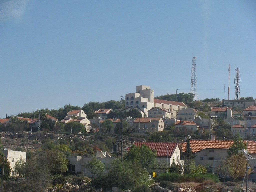 Hilly landscape with multiple houses, some with red tile roofs, a large modern building with arches, and communication towers on a sunny day.