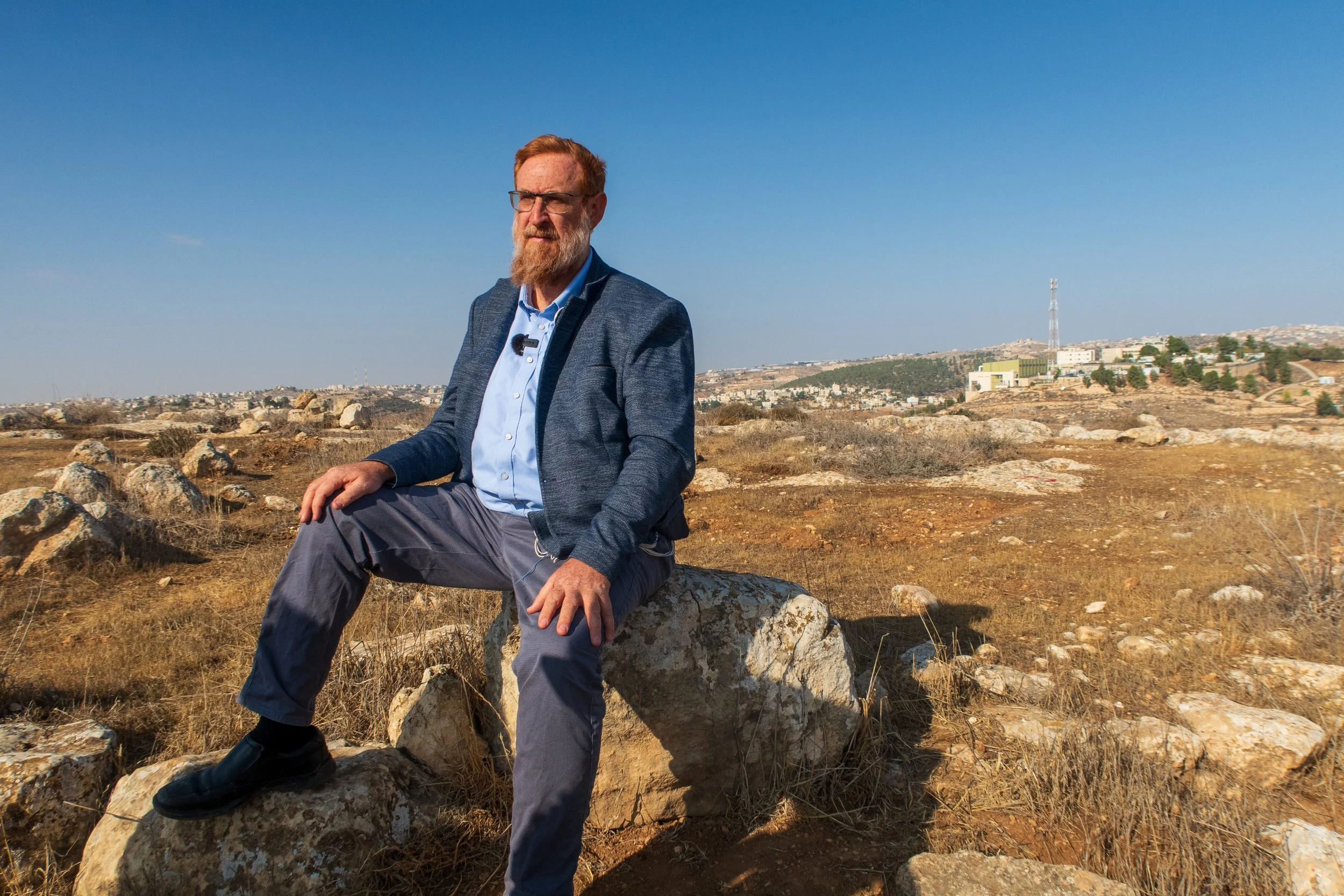 Man with glasses and beard, wearing a blue blazer and gray pants, sitting on a large rock in a dry, rocky landscape under a clear blue sky.