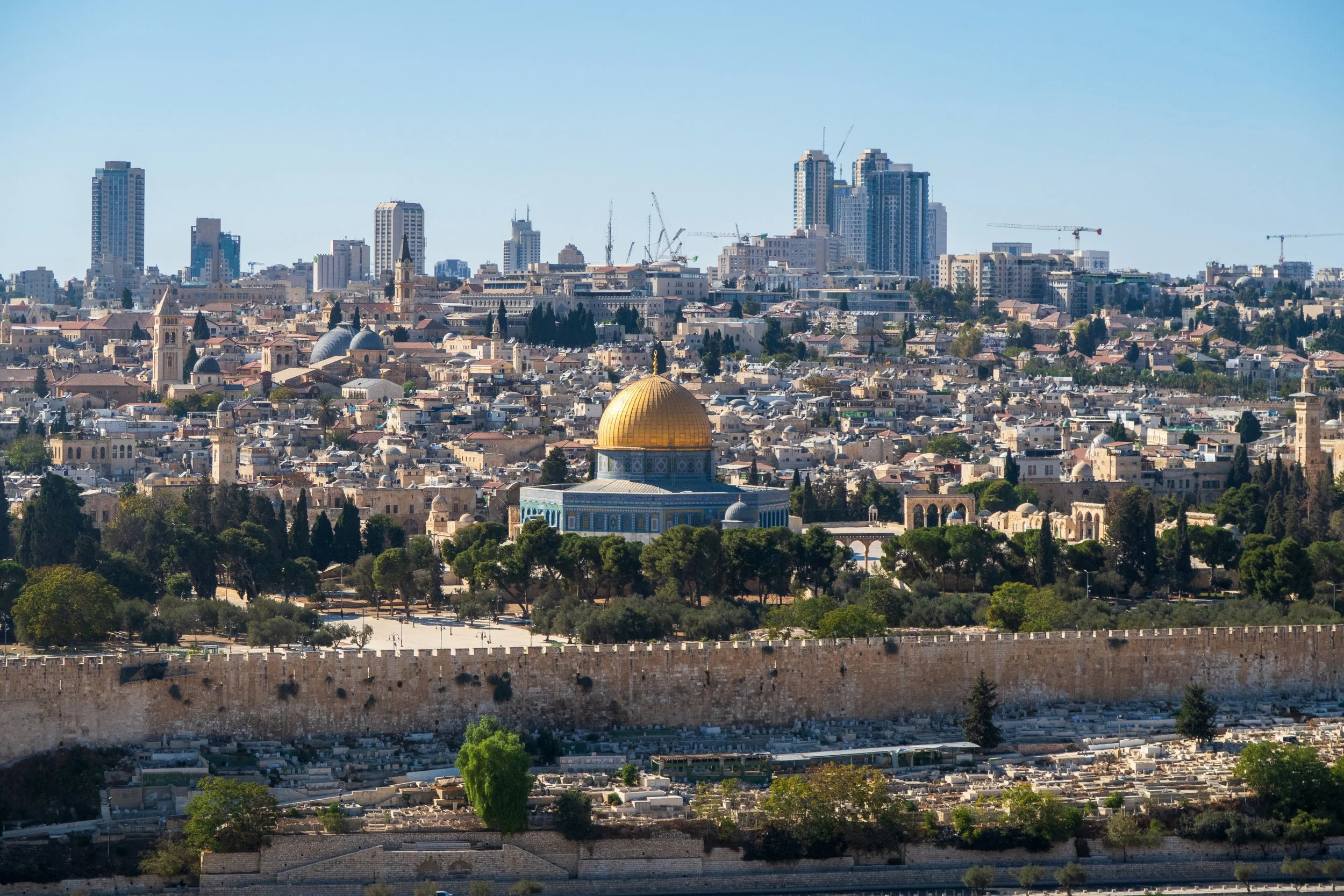 City skyline with a prominent golden-domed building in the foreground, including trees, a stone wall, and modern high-rise buildings in the background.