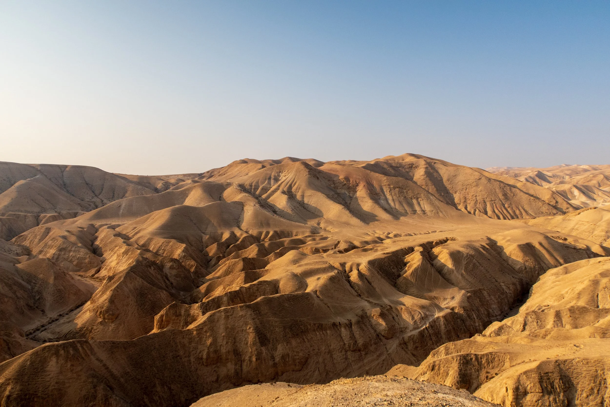 Desert landscape with sandy hills and mountain ridges under a clear blue sky.