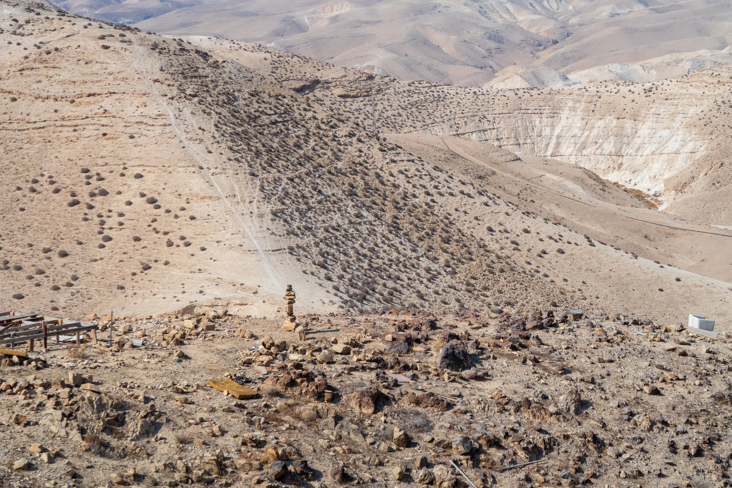 A barren desert landscape with rocky terrain, scattered rocks, and sparse desert bushes on arid hills with visible erosion patterns.