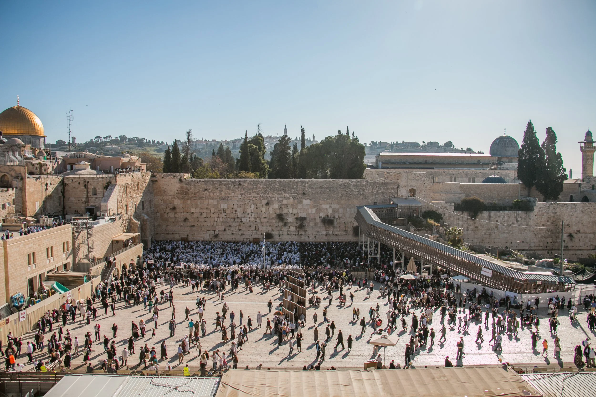 People praying and gathering at the Western Wall in Jerusalem during the day, with the golden Dome of the Rock visible on the left and other historic buildings in the background.