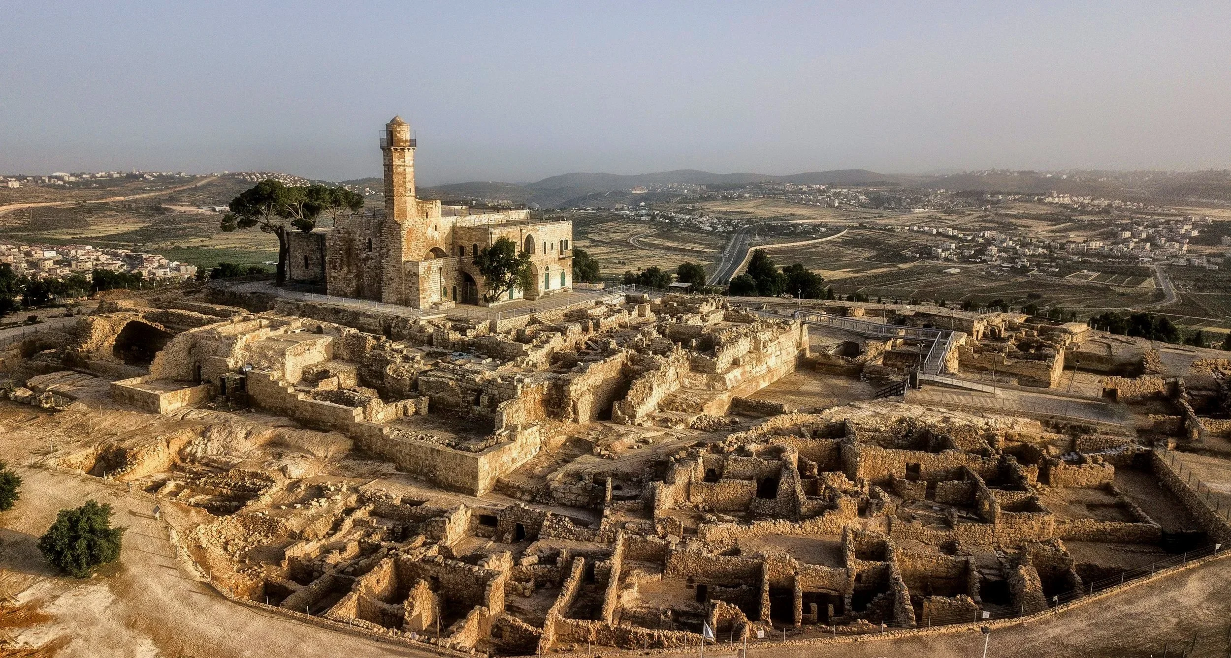 Ancient ruins with a church and a tall tower on a hill, overlooking a landscape of fields and distant buildings.