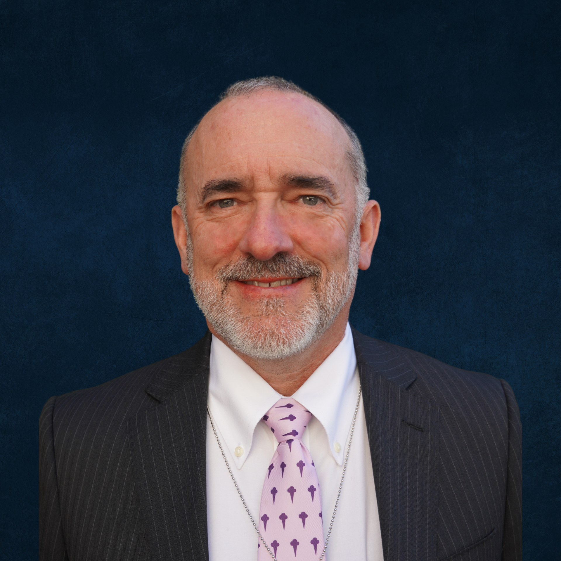 A professional headshot of a middle-aged man with a beard, wearing a pinstripe suit, white shirt, and a lavender tie with dark purple icons, against a dark blue background.