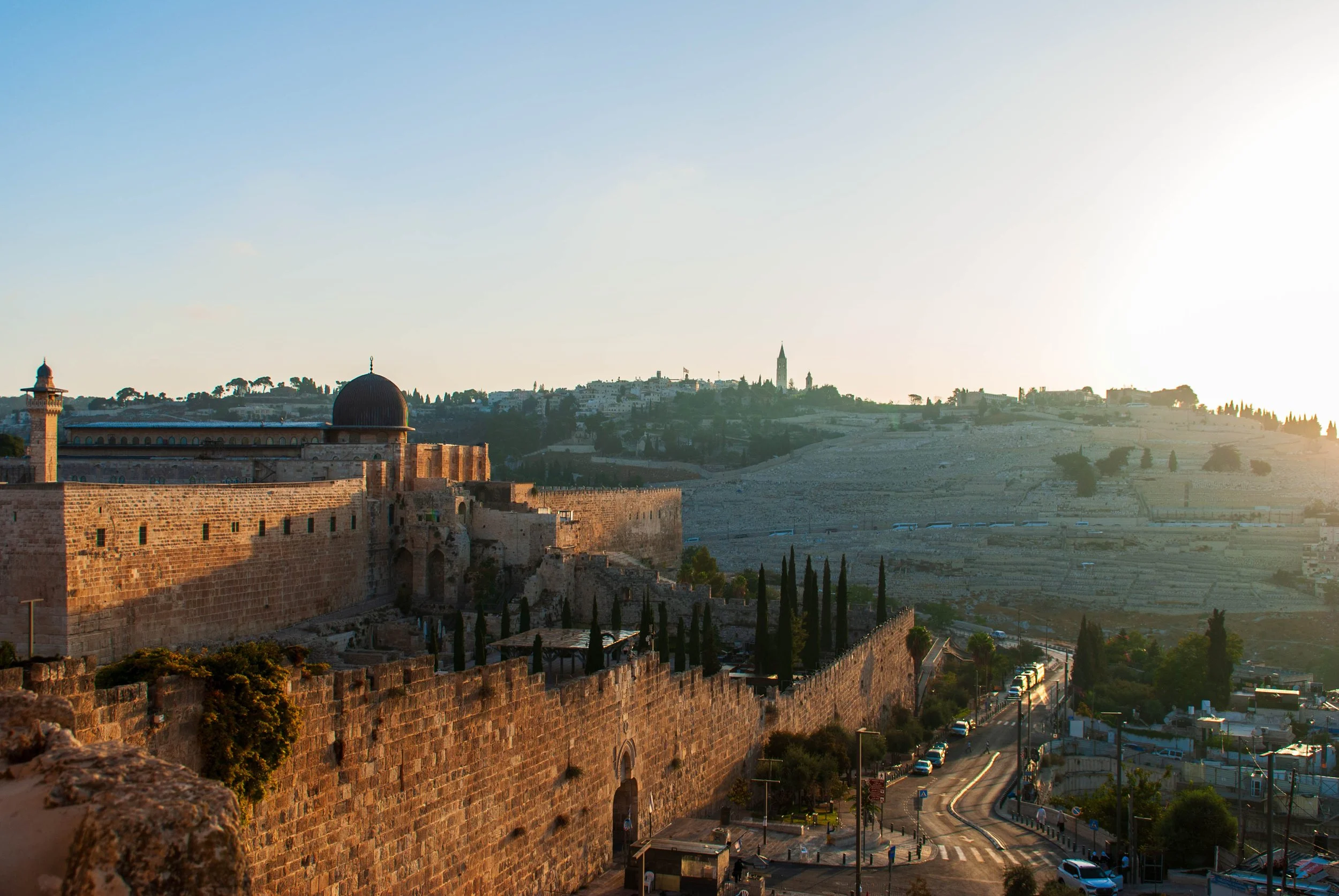 Sunrise over ancient stone city walls, domed and towered buildings, and rolling hills with sparse vegetation.
