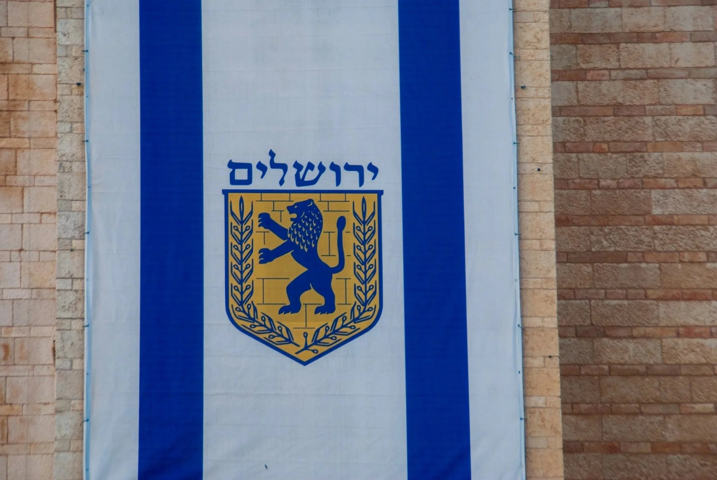 A flag with two blue vertical stripes on a white background, featuring a blue and yellow coat of arms with a lion, Hebrew text, and decorative laurel leaves, hanging on a brick wall.