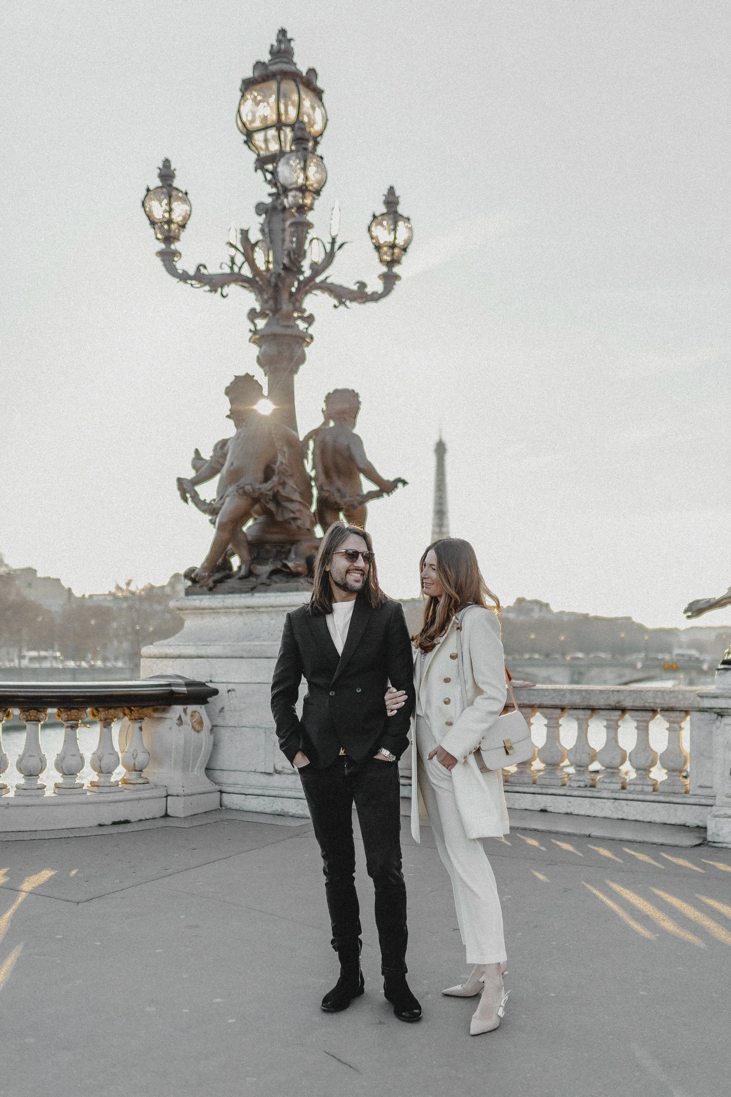 Ein Mann und eine Frau stehen Arm in Arm auf einer Brücke in Paris, im Hintergrund die Eiffel-Turm Silhouette bei Sonnenuntergang, umgeben von antiken Laternen.