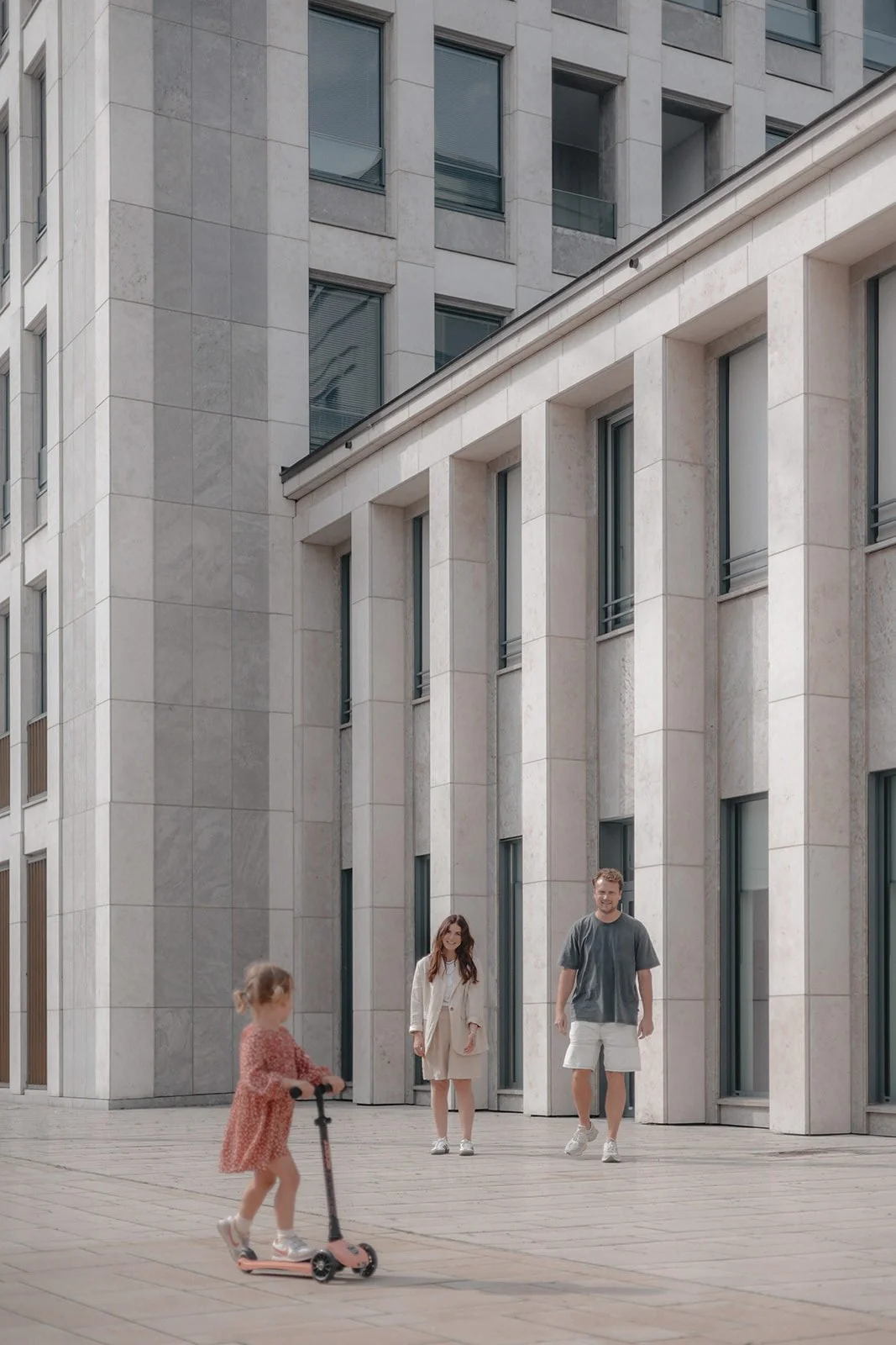 Ein kleines Mädchen fährt auf einem Tretroller vor einem modernen, hohen Gebäude mit großen Fenstern, während zwei Erwachsene, vermutlich ihre Eltern, zuschauen.